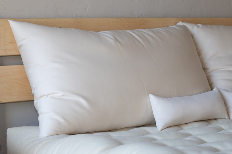 A close-up of a bed showcases large white pillows alongside a Shepherd's Dream organic wool Neck Pillow, set against a light wood headboard. The bed is completed with a luxurious quilted mattress cover, enhancing its soft and inviting appeal while promoting optimal cervical spine alignment.