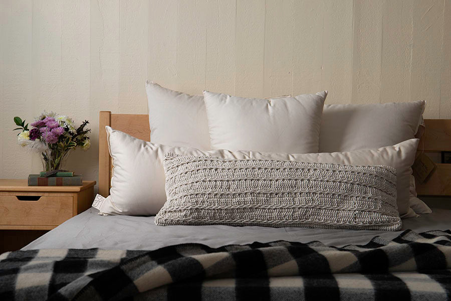 A cozy bedroom scene with a neatly arranged bed featuring white pillows, the Shepherd's Dream Wool Lumbar Pillow for back support, and a black-and-white plaid blanket. A wooden nightstand holds a vase with flowers and a stack of books.