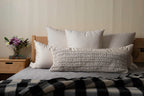 A cozy bedroom scene with a neatly arranged bed featuring white pillows, the Shepherd's Dream Wool Lumbar Pillow for back support, and a black-and-white plaid blanket. A wooden nightstand holds a vase with flowers and a stack of books.
