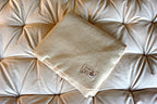 A folded beige blanket with a small white label sits on a light tufted cushion, which could be enhanced with the Baile Wool Mattress Protector for added comfort.