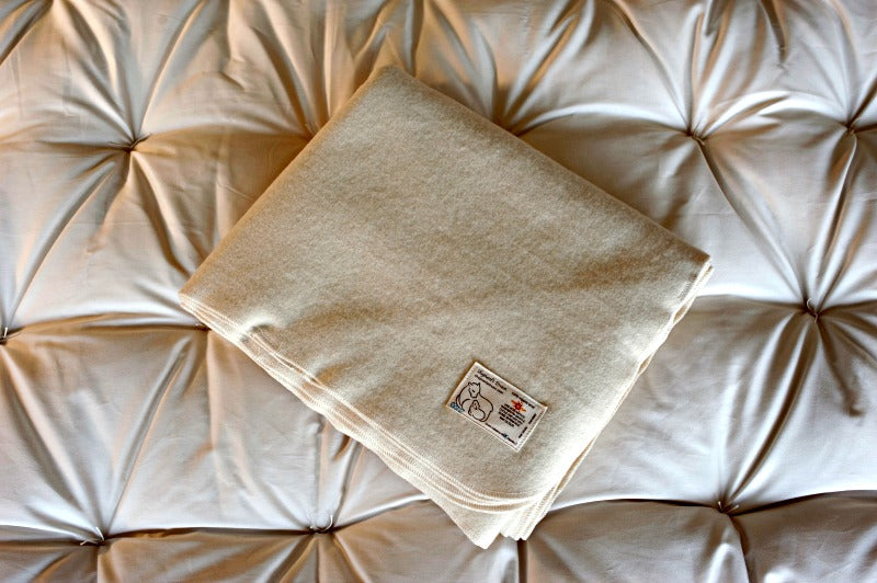 A folded beige blanket with a small white label sits on a light tufted cushion, which could be enhanced with the Baile Wool Mattress Protector for added comfort.