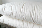 A close-up of two Ashland Organic Wool Pillows by Shepherd's Dream stacked on a wooden surface. These pillows feature a smooth texture, indicating they are clean and freshly laundered, making them ideal for a healthy bed and natural sleep environment.
