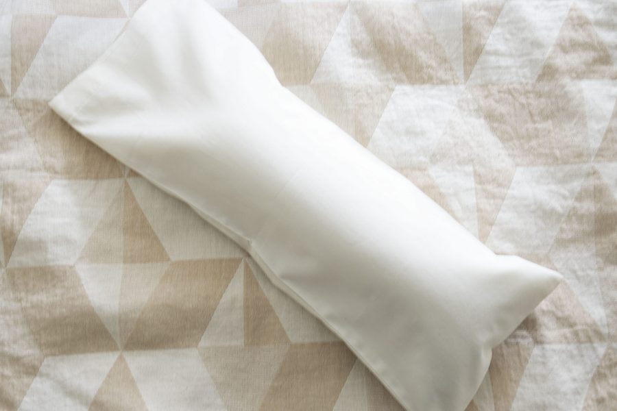 A Shepherd's Dream Neck Pillow in white, designed for cervical alignment, rests on a beige and white geometric-patterned fabric surface.