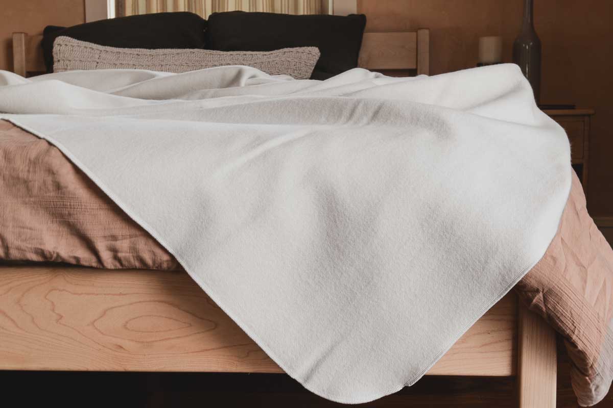 A neatly made bed with a beige blanket and pillows, topped with Shepherd's Dream Organic Merino Wool Blanket in white, draped loosely over the mattress in a cozy bedroom.