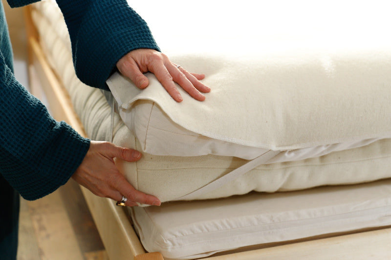 A person in a blue robe lifts the corner of a Baile Wool Mattress Protector, revealing the mattress underneath. The protector is cream-colored and washable.