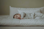 A baby sleeps peacefully on a soft, white mattress, nestled among Wool Filled Body Pillows by Shepherd's Dream. Dressed in light striped sleepwear, the child is surrounded by a serene atmosphere with a light beige background. The gentle support from the EcoWool® fill enhances the restful scene even further.