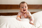 A cheerful baby dressed in a pink and white outfit is lying on the Wool Lumbar Pillow from Shepherd's Dream, which has a washable cotton sateen cover, and is propped up on its arms. In the background, there is a wooden headboard set against a light-colored wall.
