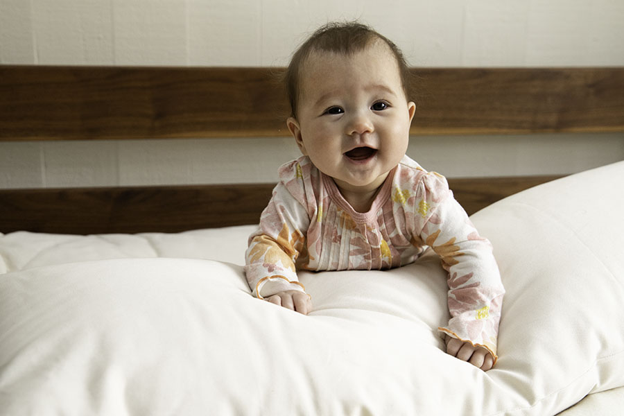 A cheerful baby dressed in a pink and white outfit is lying on the Wool Lumbar Pillow from Shepherd's Dream, which has a washable cotton sateen cover, and is propped up on its arms. In the background, there is a wooden headboard set against a light-colored wall.