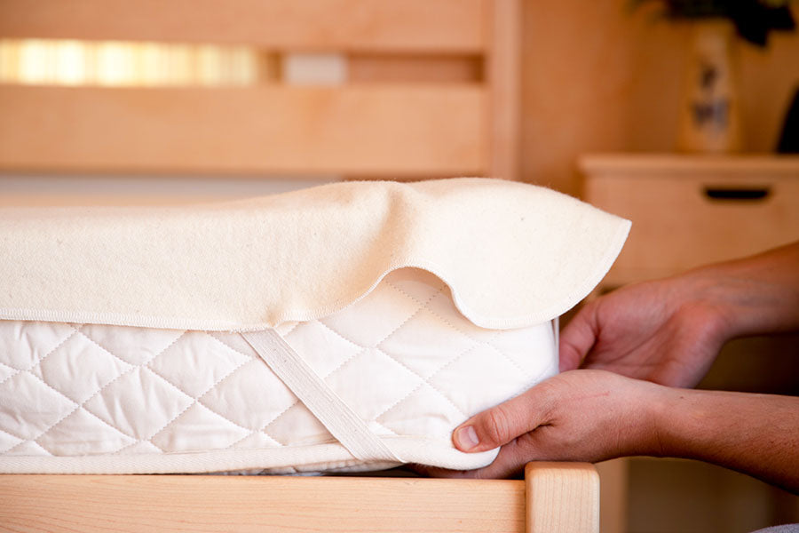 Hands fitting the Baile Wool Mattress Protector, featuring elastic straps and washable wool, onto a mattress in a wooden bed frame in a softly lit room.