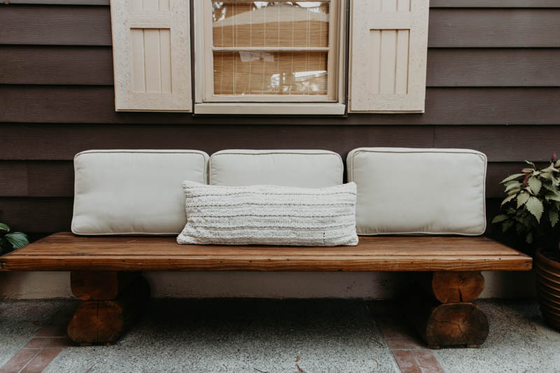 A rustic wooden bench with three white cushions is complemented by a Shepherd's Dream Wool Lumbar Pillow for added back support, resting against a brown wooden house wall. Above the bench, a window with closed shutters adds charm, and a potted plant sits beside it. The cushions feature washable cotton sateen covers.