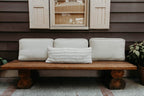 A rustic wooden bench with three white cushions is complemented by a Shepherd's Dream Wool Lumbar Pillow for added back support, resting against a brown wooden house wall. Above the bench, a window with closed shutters adds charm, and a potted plant sits beside it. The cushions feature washable cotton sateen covers.