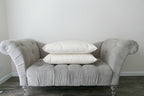 A small, gray tufted sofa with transparent legs is placed against a plain white wall. Two stacked Ashland Organic Wool Pillows by Shepherd's Dream are centered on the sofa, while the hardwood floor beneath it remains visible.