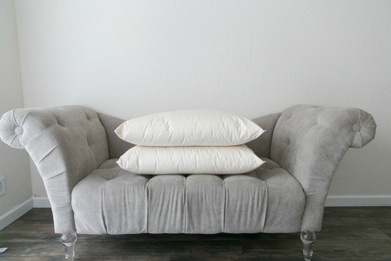 A small, gray tufted sofa with transparent legs is placed against a plain white wall. Two stacked Ashland Organic Wool Pillows by Shepherd's Dream are centered on the sofa, while the hardwood floor beneath it remains visible.