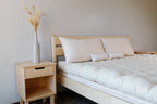 A minimalist bedroom features a light wood bed with the All Wool Mattress by Shepherd's Dream and white bedding, next to a matching nightstand topped with a tall gray vase of dried grasses. Neutral walls create a clean, simple design.
