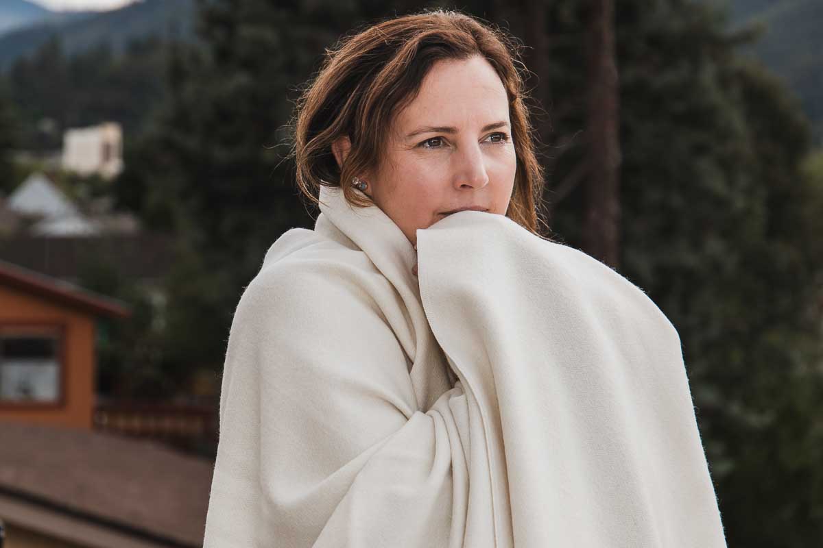 A woman stands outdoors, wrapped in a Shepherd's Dream Organic Merino Wool Blanket. She gazes thoughtfully with blurred trees, buildings, and mountains in the background on a cloudy day.