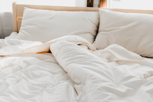 A cozy unmade bed with soft white linens, two large pillows, and sunlight streaming through a window features the Ashland Organic Wool Comforter by Shepherd's Dream. The comforter has a slightly ruffled look that makes it appear comfortable and inviting.