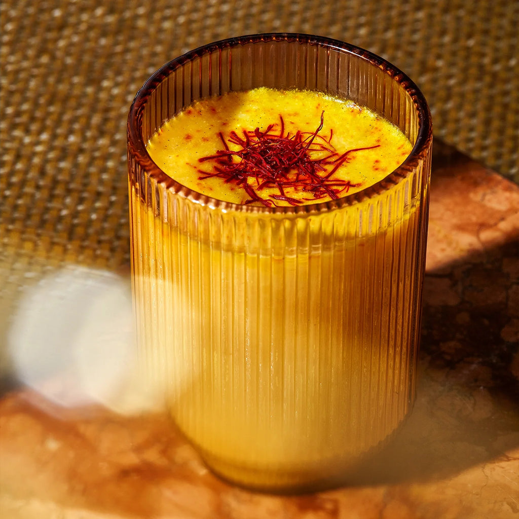 A ridged glass filled with a golden yellow saffron latte, topped with delicate saffron strands, sits on a warm-toned marbled surface. The serene atmosphere is enriched by the deep hues and textured woven backdrop, making it perfect for enhancing mood and promoting restful sleep—a hallmark of THE FULLEST's Warm Feelings 30-Day Supply.