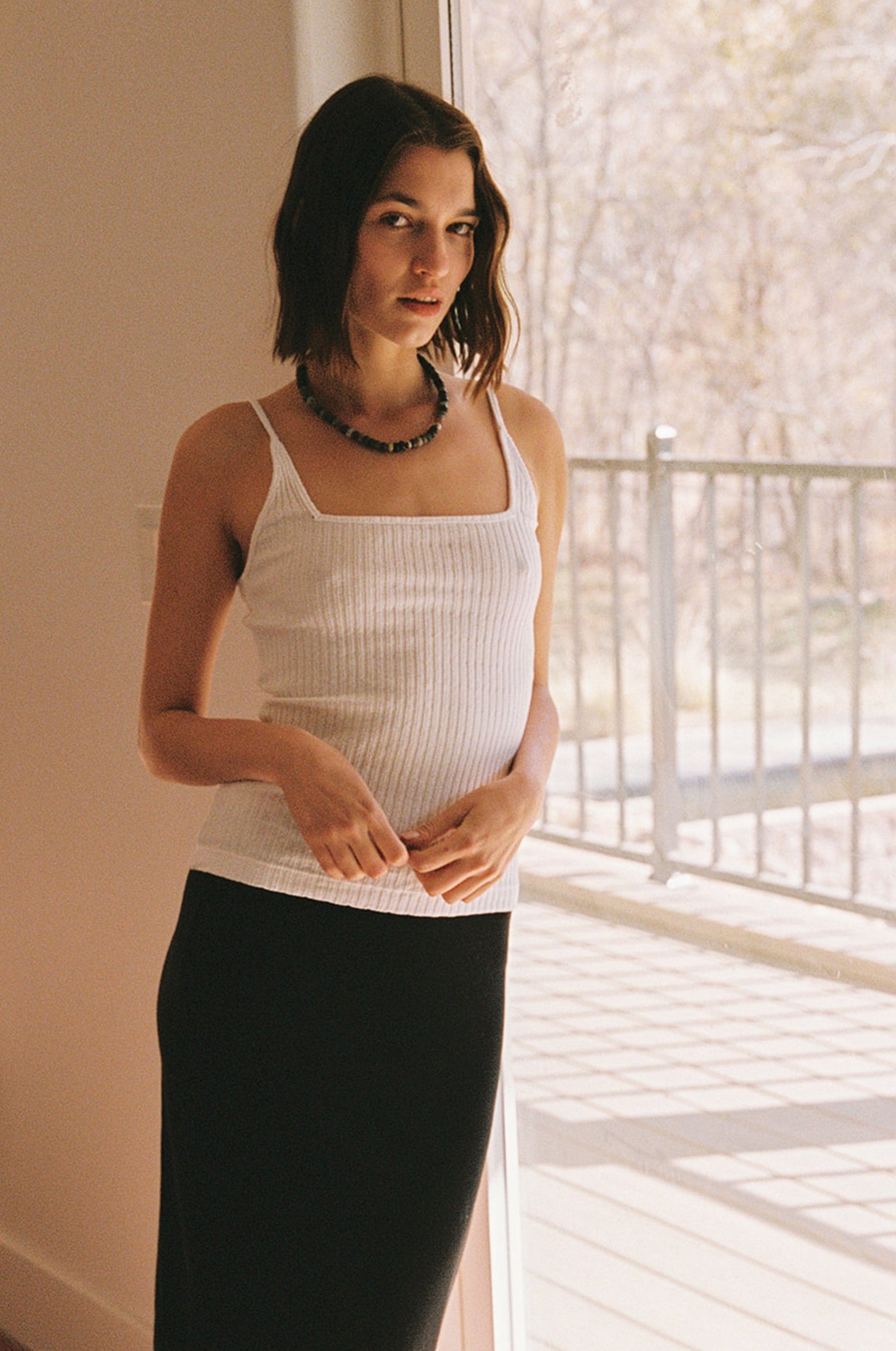 A woman with shoulder-length brown hair wears the LA Relaxed Pointelle Square Cami, paired with a black skirt and beaded necklace, standing indoors by a sunlit glass door with an outdoor railing visible.