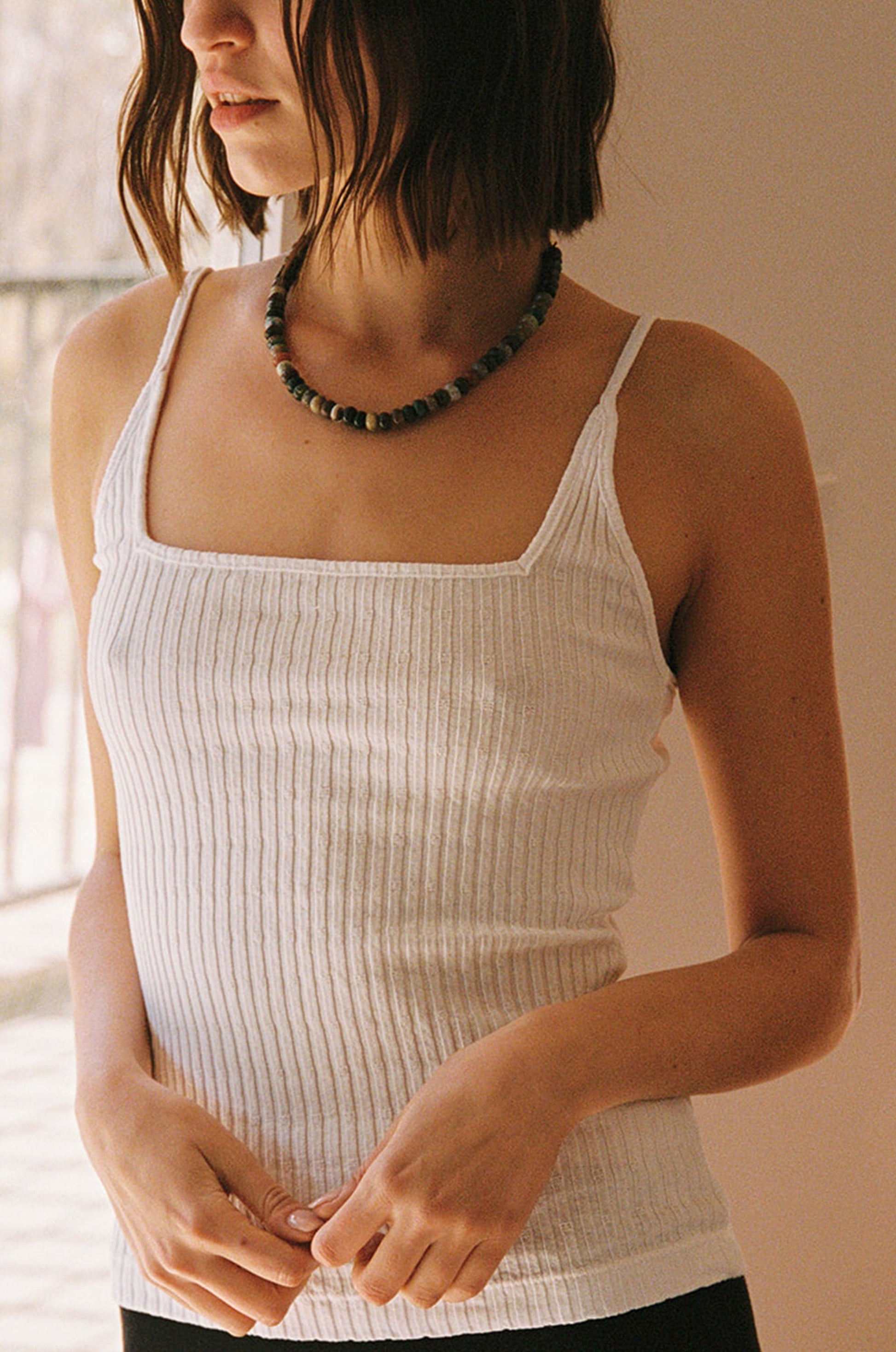 A woman with shoulder-length brown hair wears the LA Relaxed Pointelle Square Cami and a beaded necklace, standing indoors in soft natural light. Her face is partially out of the frame.