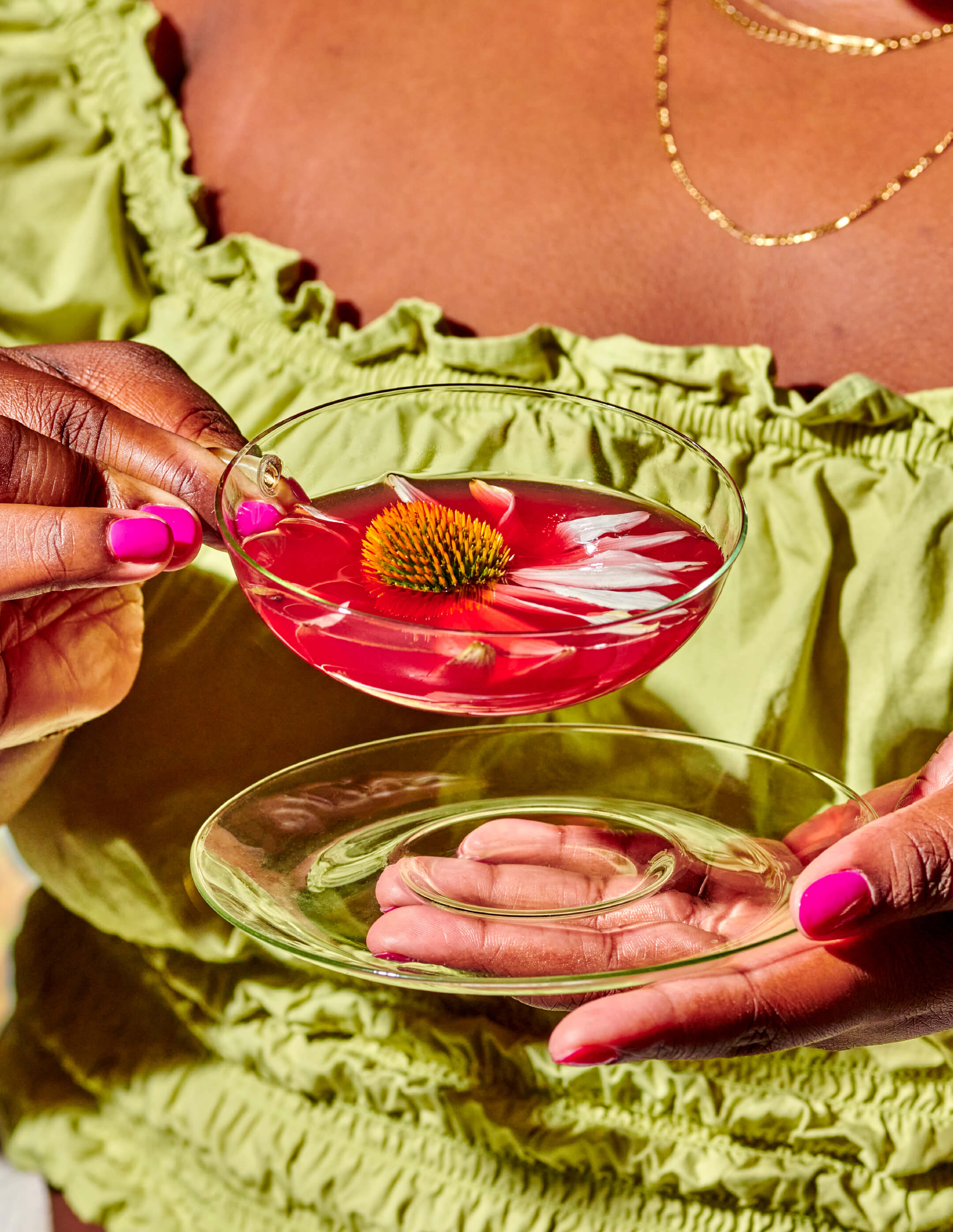 A person in a green ruffled top, with a gold necklace, holds a glass saucer and a clear glass cup filled with the pink immune toddy beverage. Inside the drink is a vibrant orange and green flower, highlighting the Lemon-Ginger Immune Toddy Sachets by WOODEN SPOON HERBS. Their nails are painted pink.